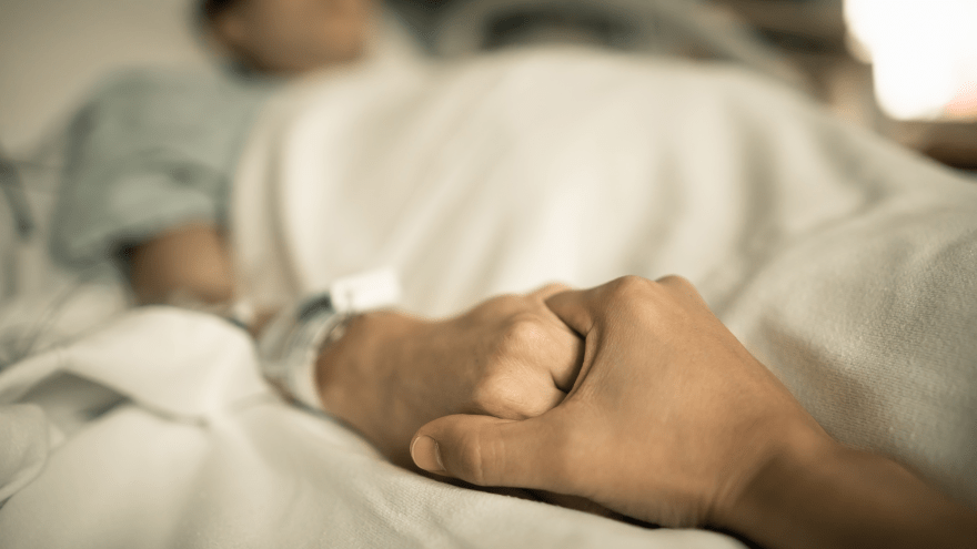 eine Hand hält die Hand einer in einem Krankenbett liegenden Person